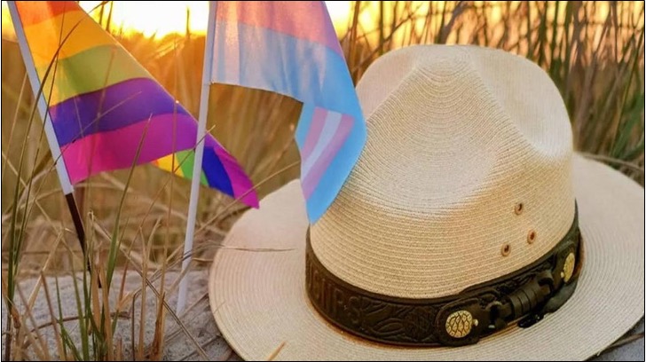 A color photograph of a NPS ranger hat sitting on a sand dune at sunset with an LGBTQ Pride flag and a transgender pride flag flying next to it.
