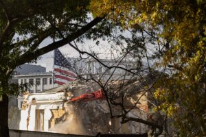 A color photograph showing a building demolition. In the background is a US flag and the US White House.