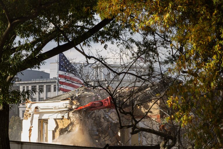 A color photograph showing a building demolition. In the background is a US flag and the US White House.